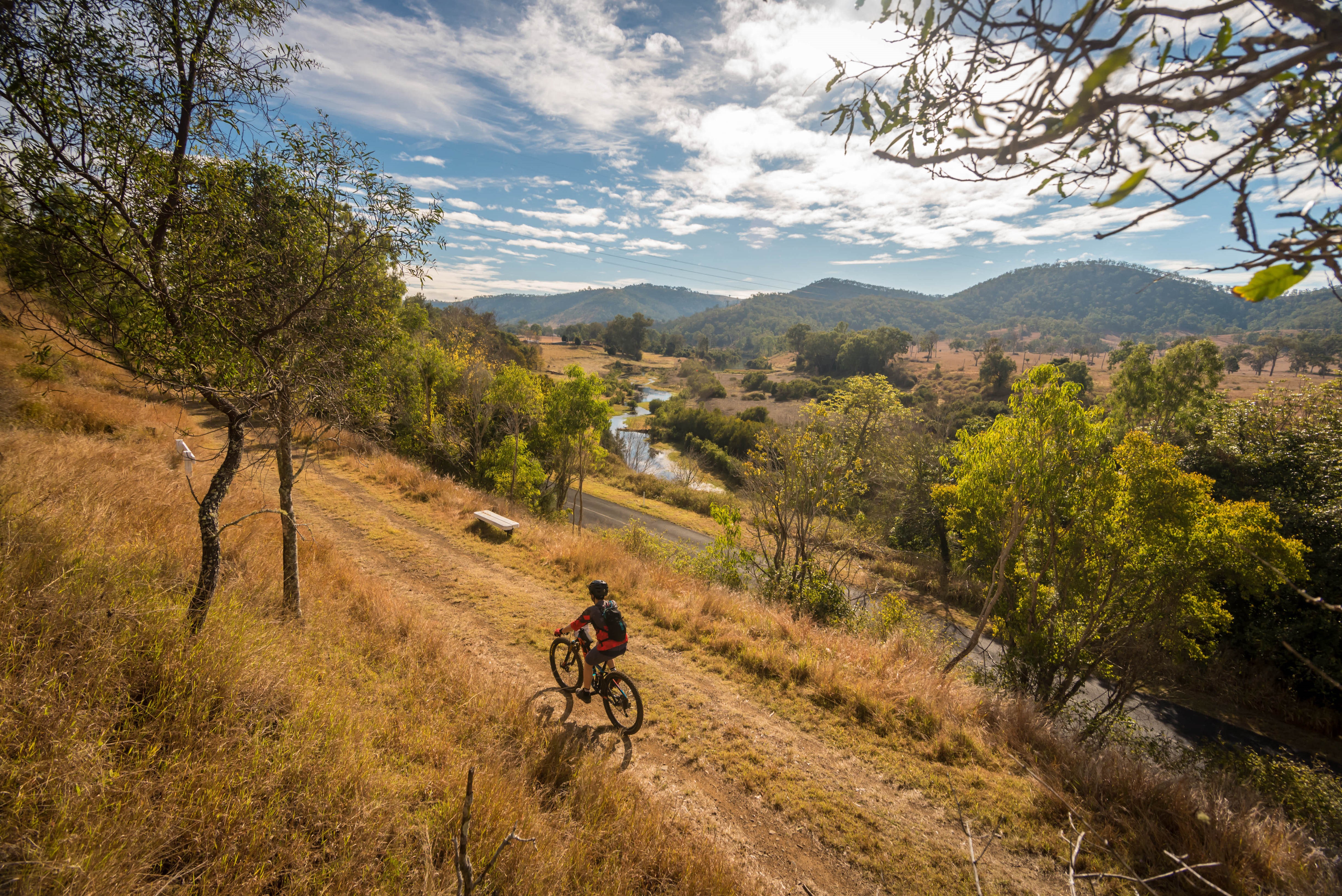 A bicycle rider on a plain, surrounded by trees with cloudy sky and water visible below