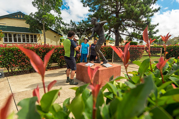 A statue of a tennis player in front of 2 buildings, surrounded by trees