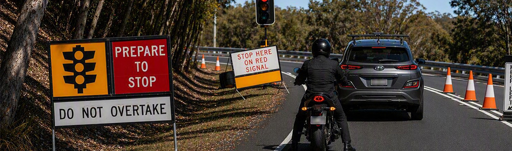 Traffic signs on the side of the road and a red light camera with a car and a person on a motorcycle waiting for it to turn green and traffic cones in the middle of the road.
