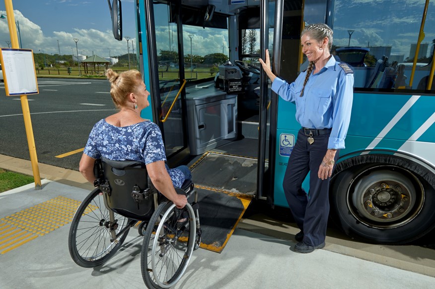 Bus driver assisting a person in a wheel chair to board a blue bus at a bus station.