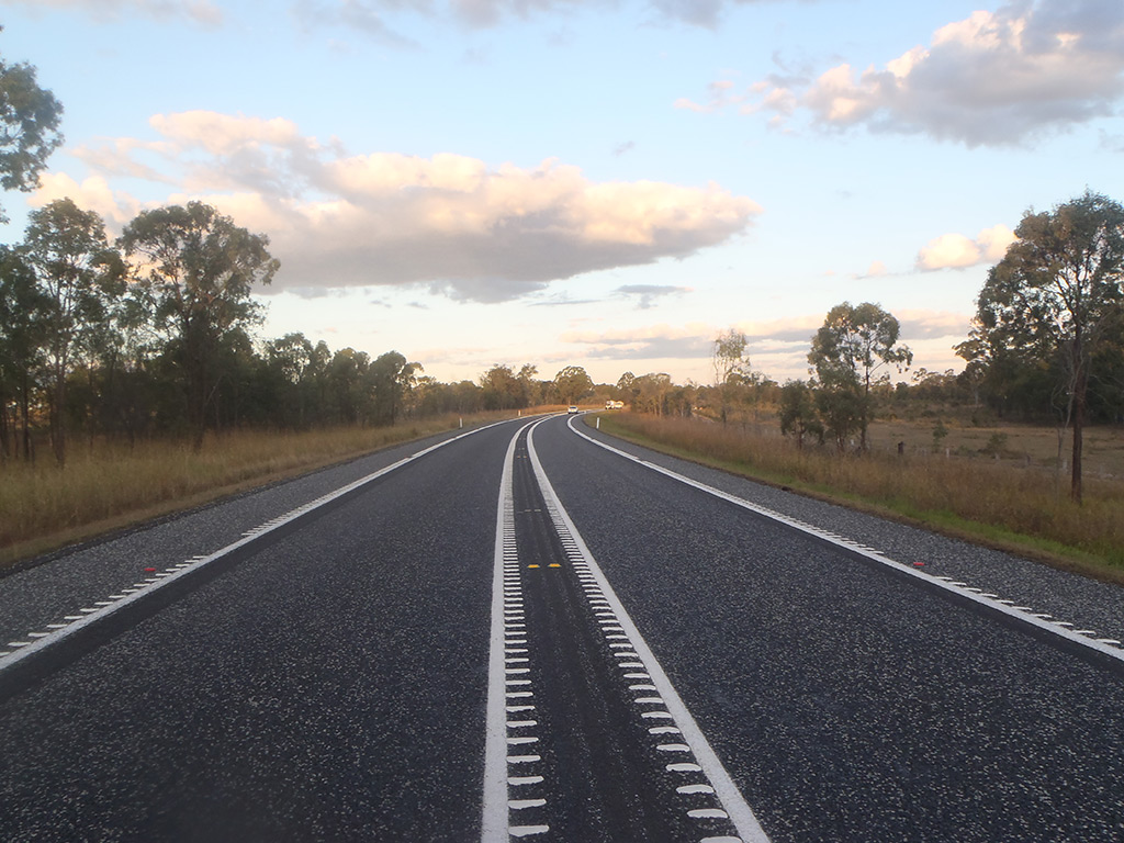 A single-lane highway with one lane in each direction, featuring a wide centre line treatment as part of the Bruce Highway Targeted Safety Program. The road is surrounded by bushland and set against a blue sky with scattered clouds.