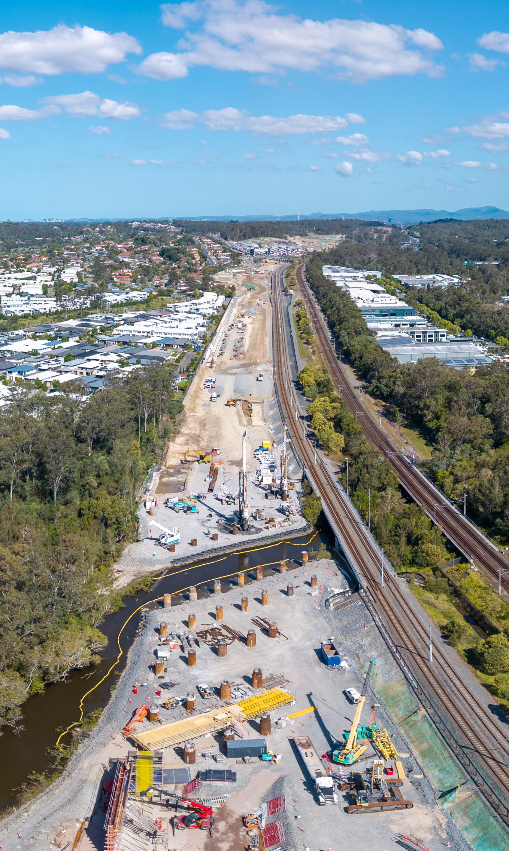 Construction site of the works happening at Coombabah Creek looking south with building and trees on either side and two sets of train tracks on the right-hand side of the construction site. 