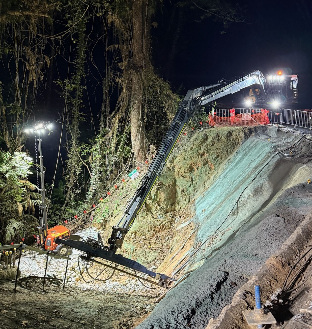 Nightwork drilling of the downslope side of the Mossman - Mount Molloy road with lights at the bottom of the slope and trees in the background.