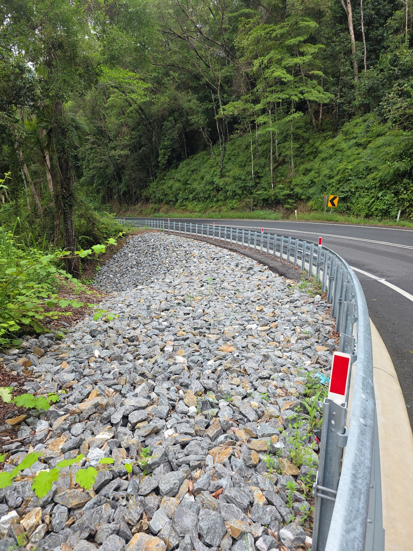 Rocky side of the road that curves left with a guard rail and trees on either side of the road.