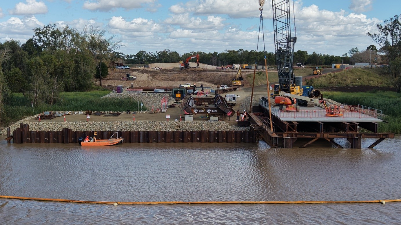 Front view of the temporary bridge work platform installation on a river with trees and a cloudy sky in the background. 