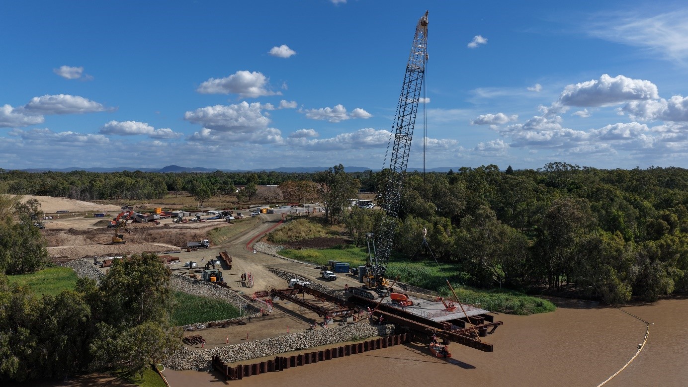 Aerial view of construction work with a crane on a riverbank with trees around the construction area and a cloudy blue sky in the background.