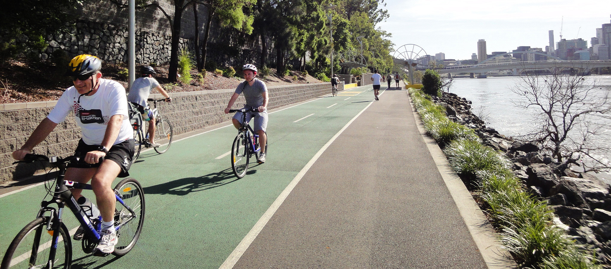 Image showing bike riders, joggers and walkers using the Bicentennial Bikeway, beside the Brisbane River with Brisbane city skyline in background.