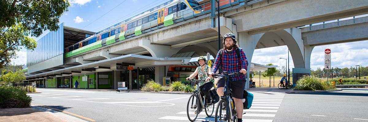 2 people cross a pedestrian crossing on bikes in front of a train station