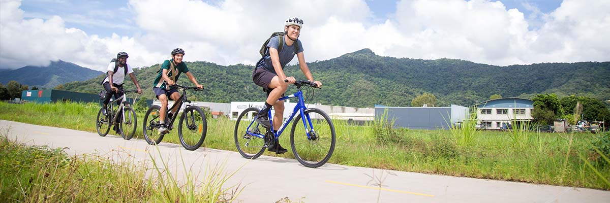 3 bike riders riding in single-file on a concrete path in a grassy area with mountains and some buildings in the background.