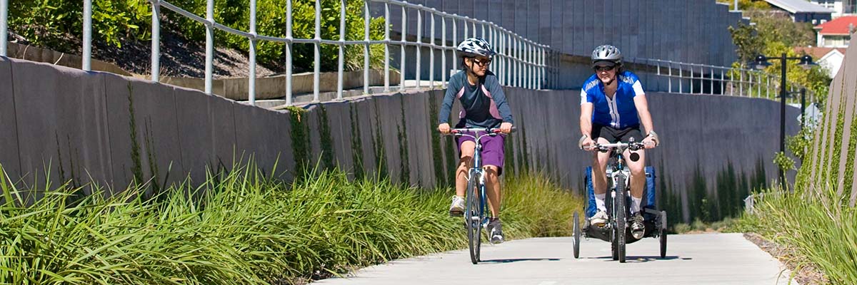 2 people riding bikes on a slightly uphill path through a cutting. One bike is towing a small trailer.