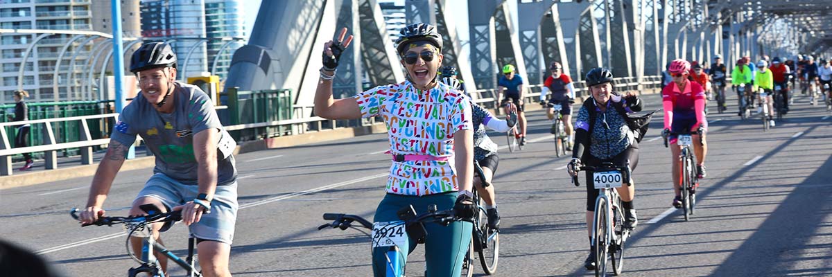 A crowd of bike riders with numbers attached to the front of their bikes riding across a multi-lane bridge with no cars.