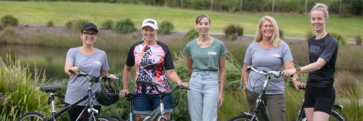 5 women stand with 3 bikes in a large outdoor space near a still body of water.