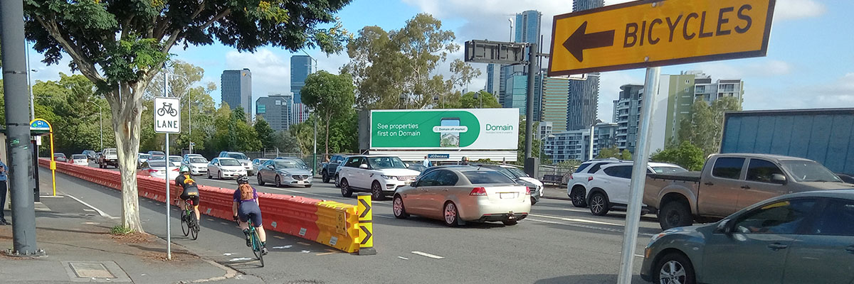A street full of cars with a city skyline in the background. A roadwork barrier is set up between the car lanes and a bicycle lane, which 2 bike riders are riding on. A yellow sign in the foreground says "bicycles" with an arrow pointing in the direction the bike riders are riding.