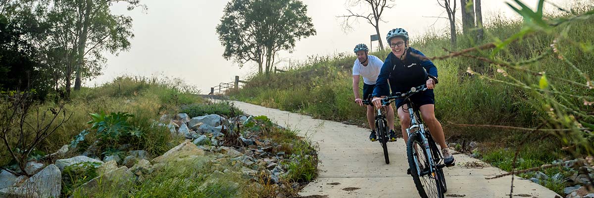 2 bike riders riding downhill on a concrete path between rocks, trees and long grass.