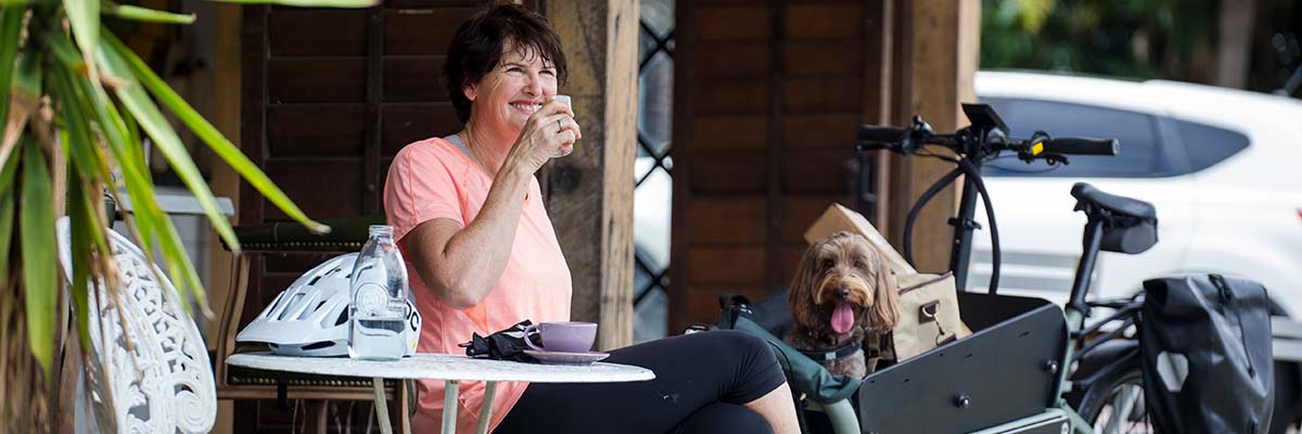 A smiling person in activewear sitting at a small outdoor table with a mug of coffee and holding a glass of water. There is a bike helmet and set of gloves on the table. A modified bike is parked to the side with a crate at the front holding a small dog and some bags.