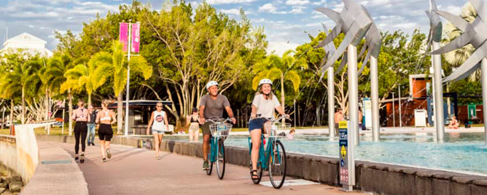 A child, and adult and a teenager ride bikes on a path next to an outer-city road. All 3 wear helmets and the child and adult wear gloves.