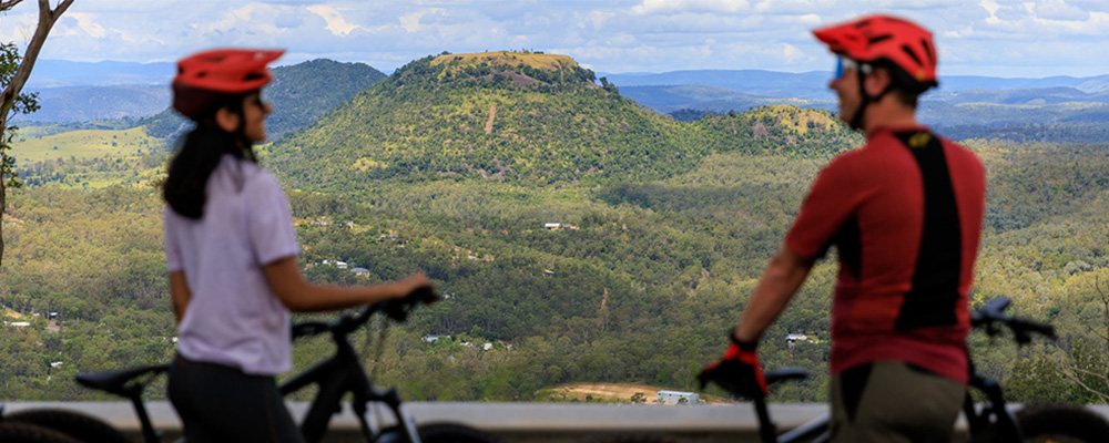 A child, and adult and a teenager ride bikes on a path next to an outer-city road. All 3 wear helmets and the child and adult wear gloves.