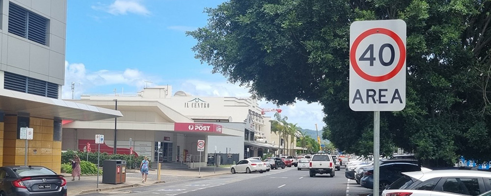 A street with buildings, trees, people walking, cars driving, cars parked in parking spots, 40km/hour speed limit painted in white on the road, and 40km/hour speed limit and parking signs.