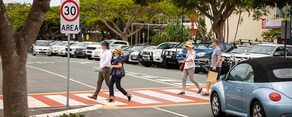 People walking across a red and white zebra crossing, with a blue car at a stop waiting for them to pass. There are trees, building and parked cars in the background. 