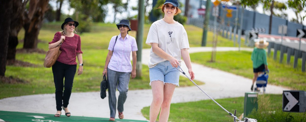 A person walking a dog on a leash at a local park.  Behind her, two people are talking with each other as they walk. 
