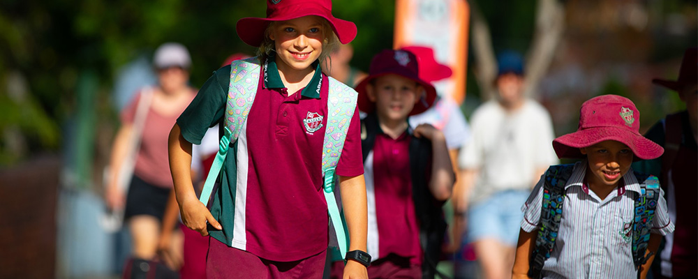 A group of children smiling, wearing uniforms, hats and carrying backpacks walking to school.
