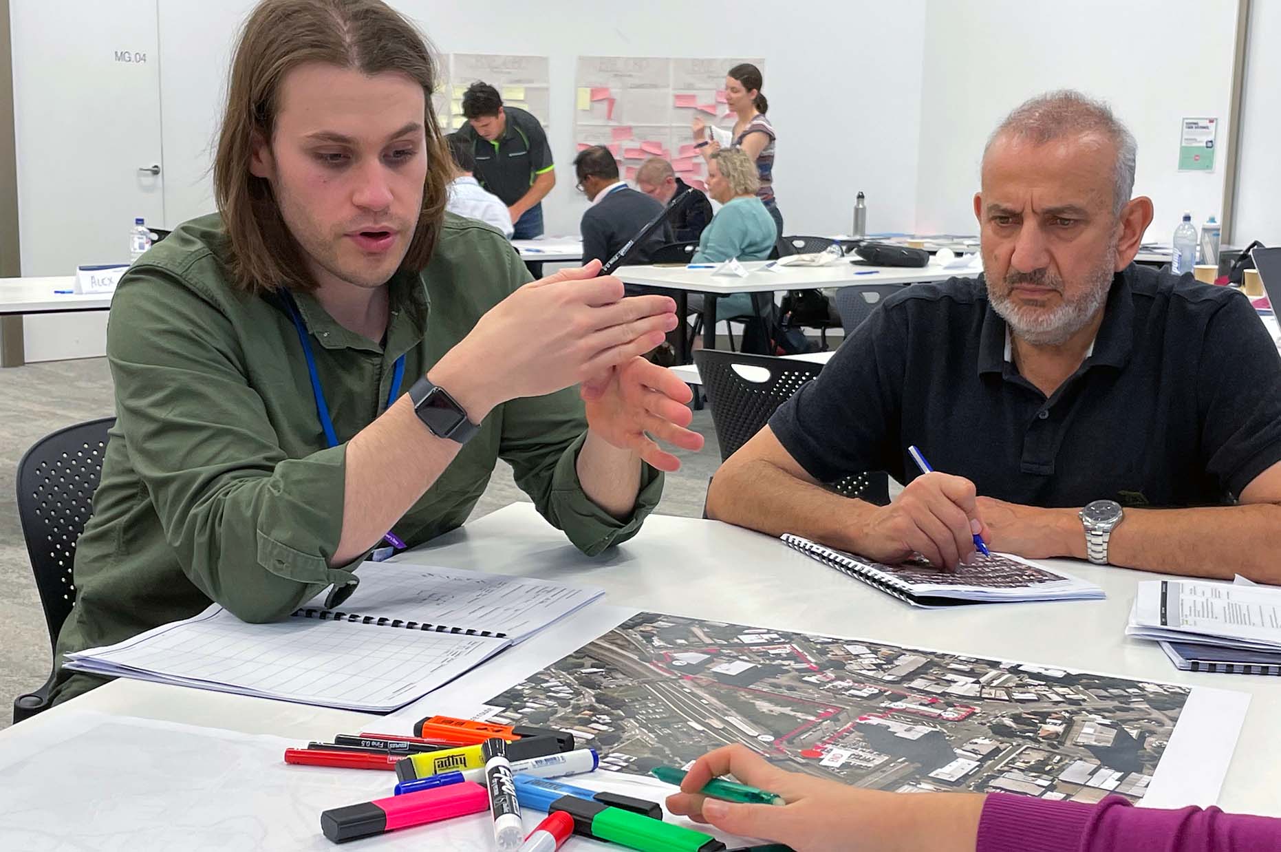 two people sitting down at a table with a map, one person is taking notes, the other is talking